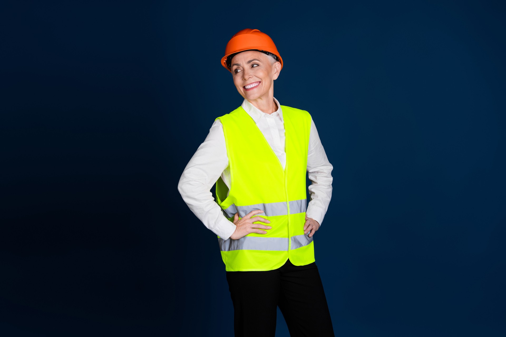 Confident middle-aged businesswoman in a high-visibility vest and orange hardhat, stands on a dark blue background